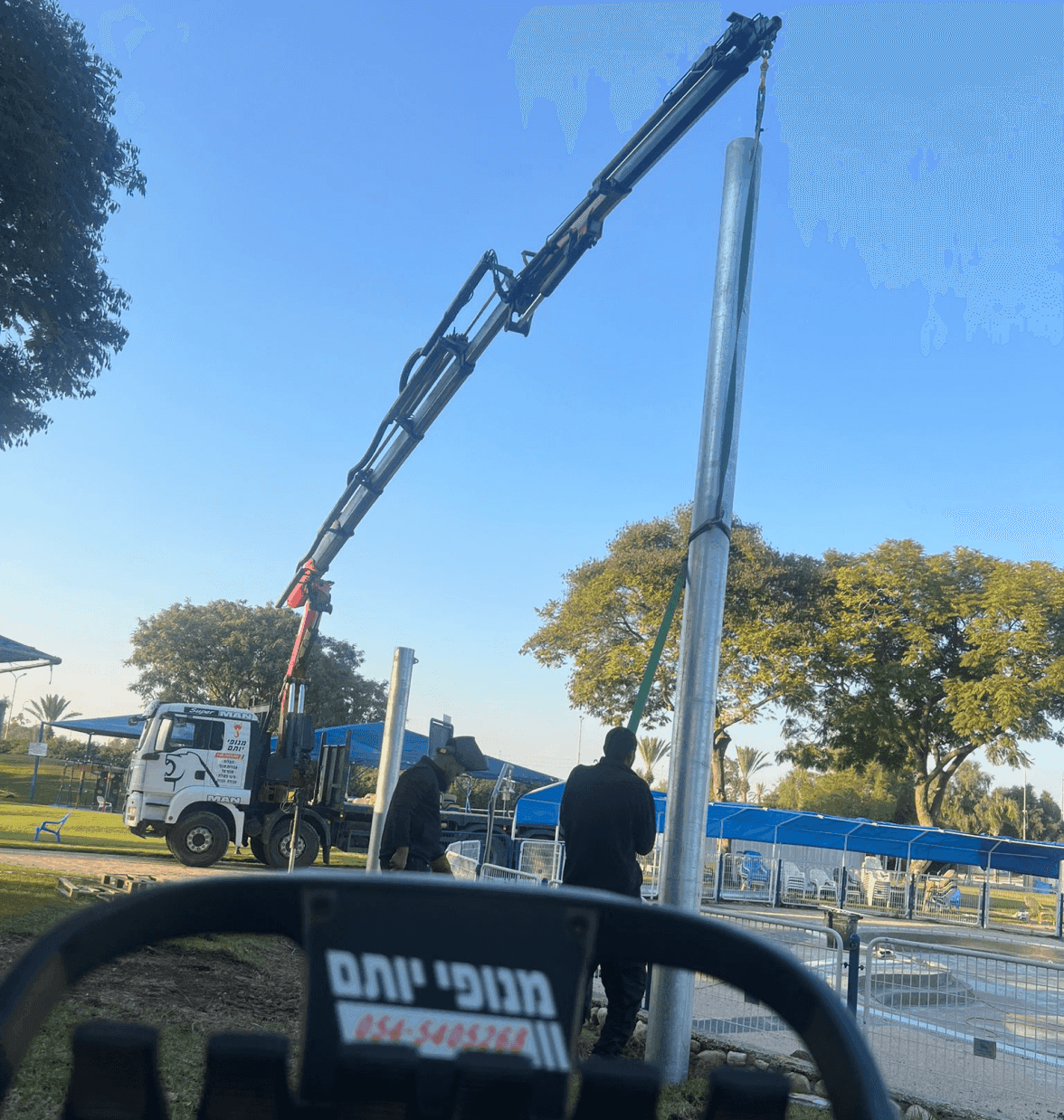 עמוד מתכת תלוי ברצועת הנפה לצד משאית שעליה מופיע השם מנופי יותם